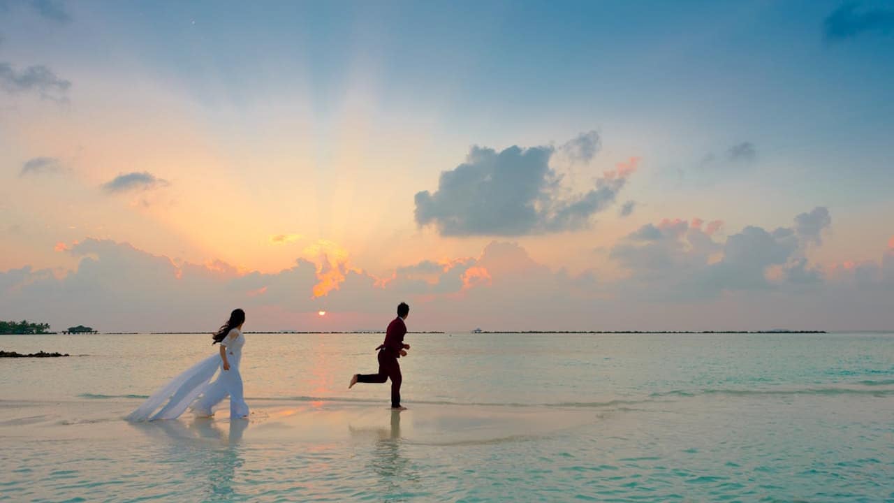 A bride in a white dress and a groom are running along the edge of the ocean at sunset.