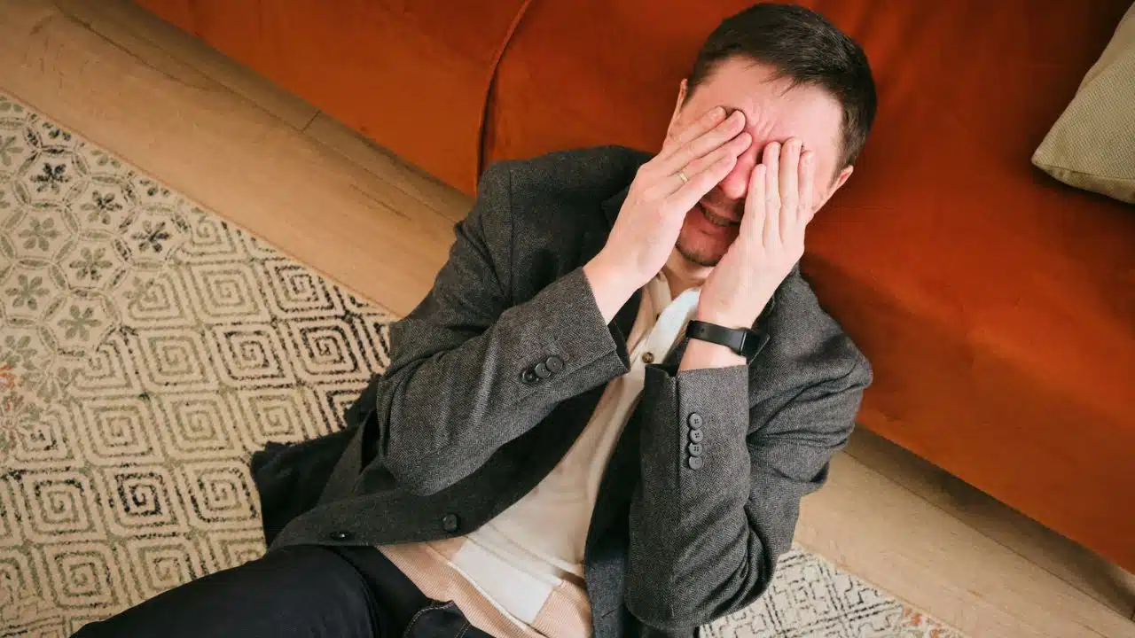 A man in a gray jacket lies on a patterned rug, covering his eyes with his hands.