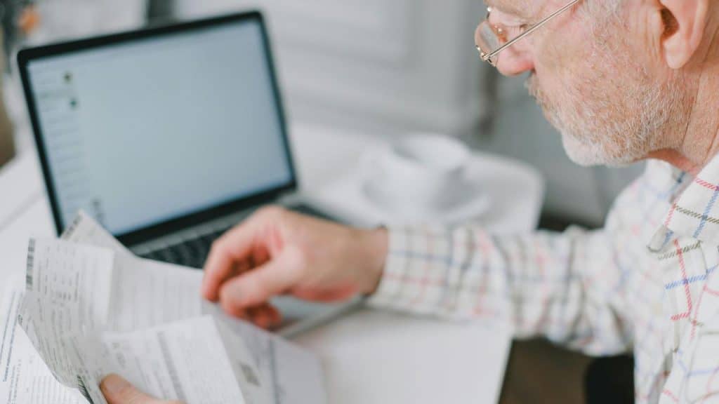 A senior man reviewing papers beside a laptop.