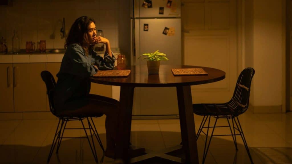 Woman sitting alone at a table in a dimly lit room, with an empty chair.
