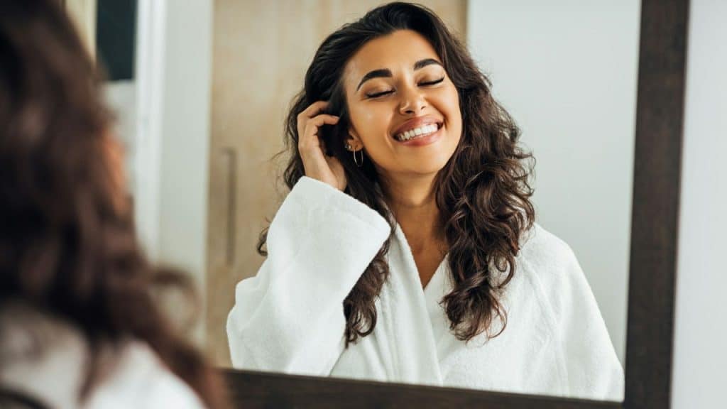 A woman in a white robe happily touches her hair, looking into a bathroom mirror.