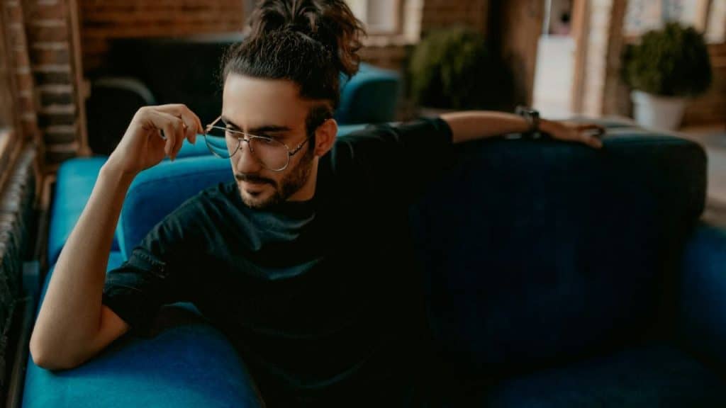 Man with bun and glasses sitting on a blue couch, hand near his face.