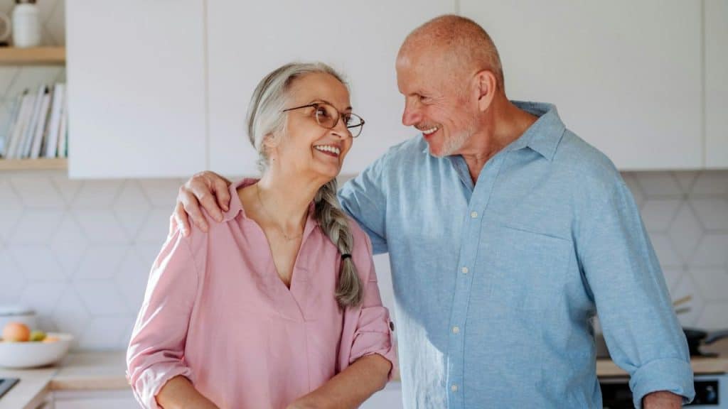 An older couple smiles at each other, standing in a bright kitchen with arms around her.