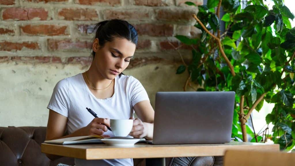 Woman working at a table, writing in a notebook next to a laptop and a mug.