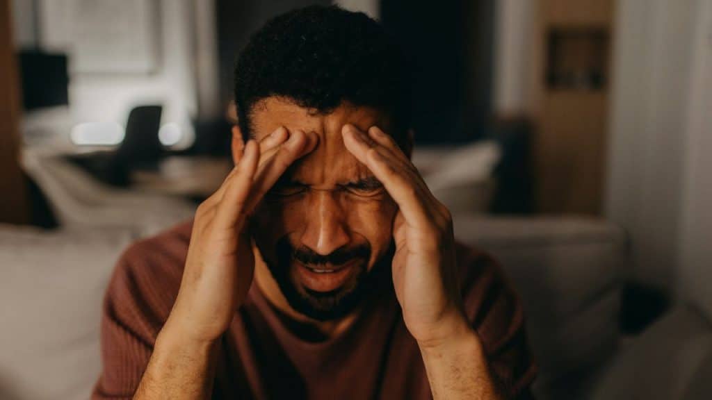 A distressed Black man in a reddish-brown shirt with hands pressed to his temples.