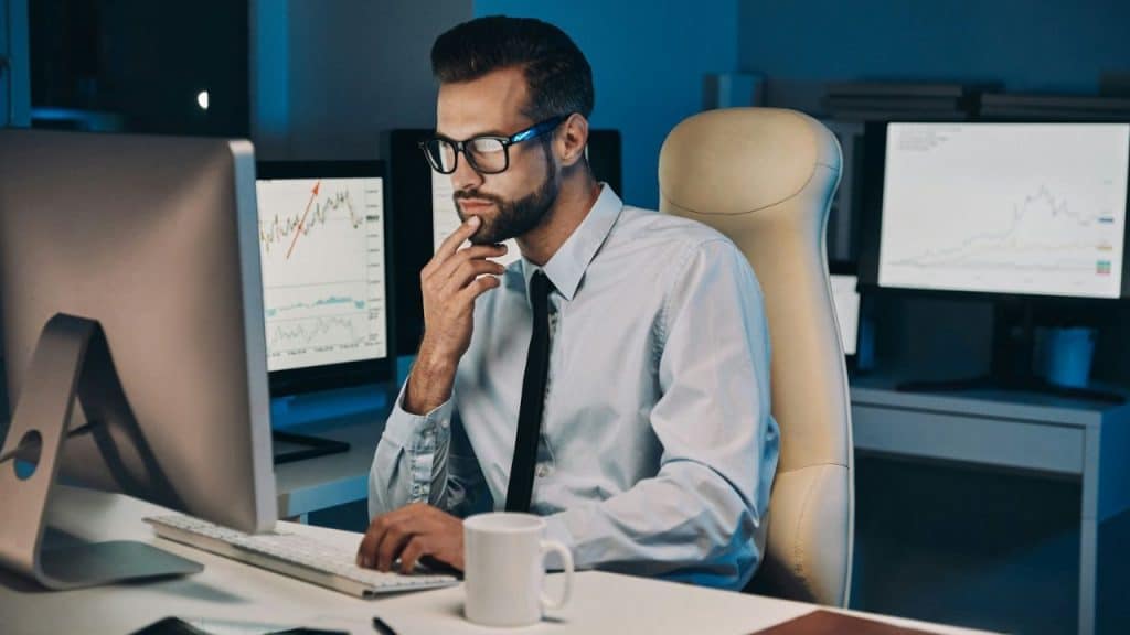 A serious man works at a desk, looking at multiple computer monitors with charts.