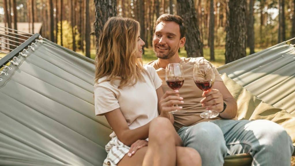 A couple in a hammock in a forest holding up two glasses of red wine.