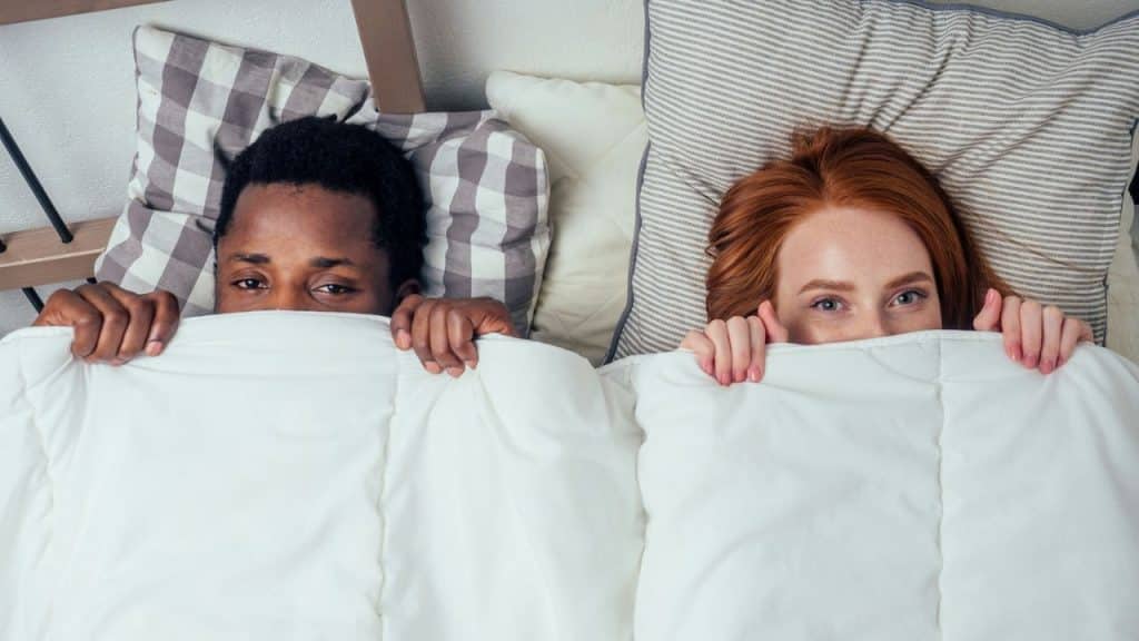 A man and a woman lie in bed, pulling a white blanket up to cover their noses.