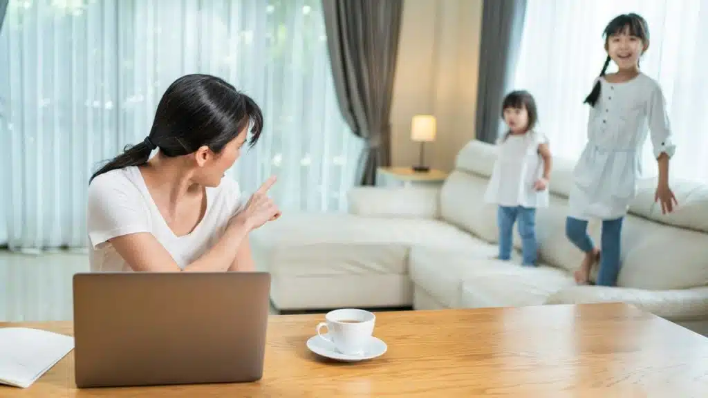 A mother working on a laptop sternly points at her two young children playing on a couch.