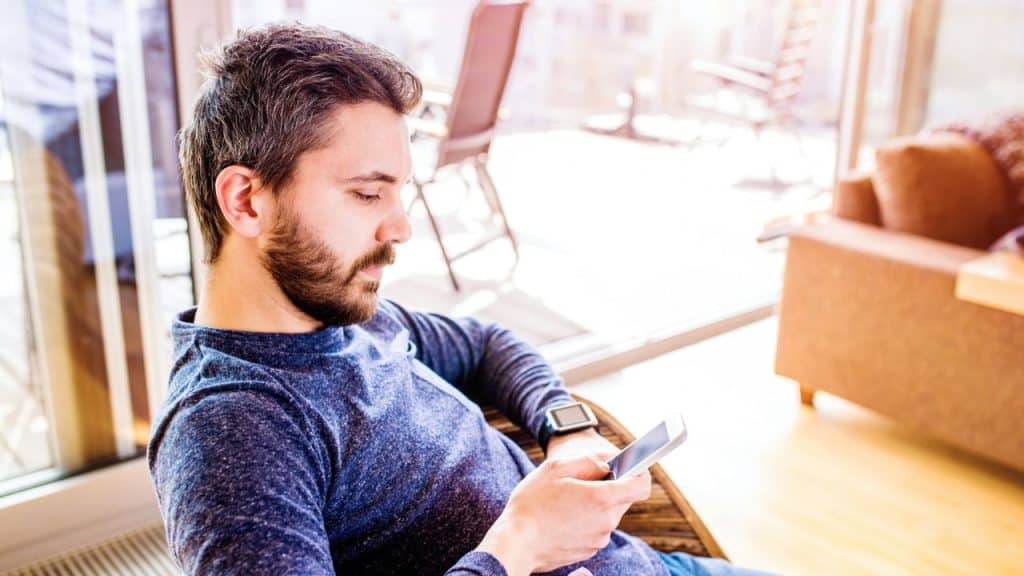 A bearded man in a blue shirt sits indoors, looking down intently at his smartphone.
