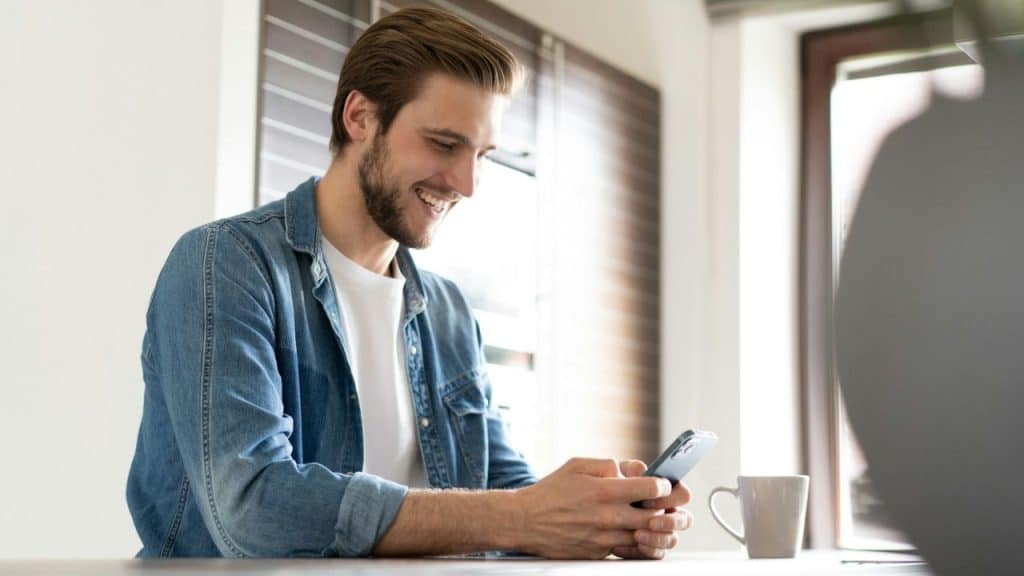 A smiling, bearded man in a denim shirt looks at his smartphone next to a coffee mug.