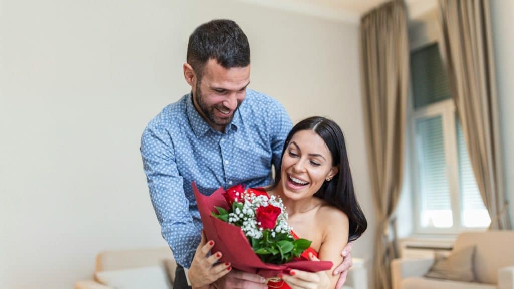 A man smiles while presenting a bouquet of red roses to a surprised and delighted woman in an indoor setting.