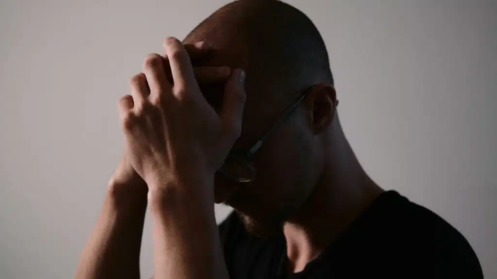 Man in dark clothing and glasses sits with hands clasped over his forehead in shadow.
