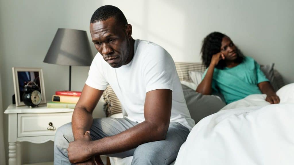 A distressed man sits on a bed while a woman sits behind him, both looking away.