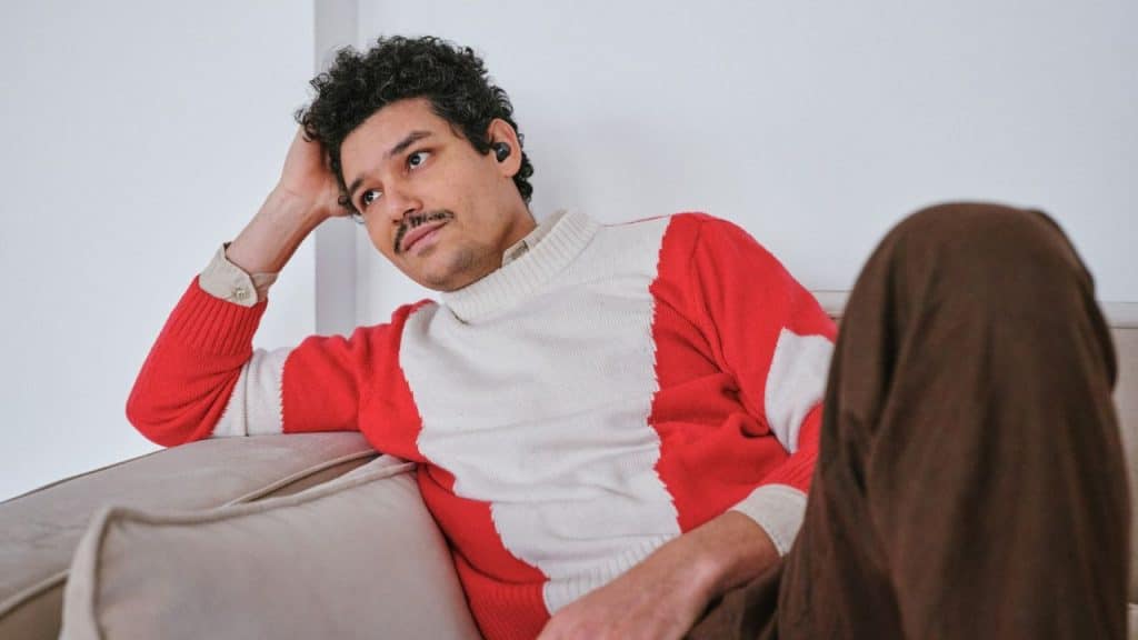 A man in a red and white sweater is sitting on a sofa looking thoughtful.