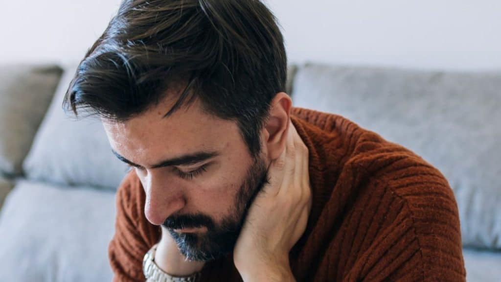 A man with dark hair and a beard is sitting, holding his hand to his neck.