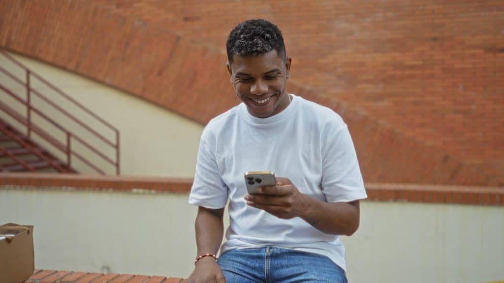 Smiling man in a white t-shirt and jeans using his smartphone outdoors.