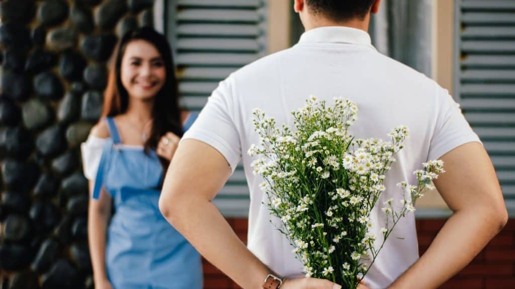 A man holding a bouquet of white flowers behind his back, surprising a smiling woman.