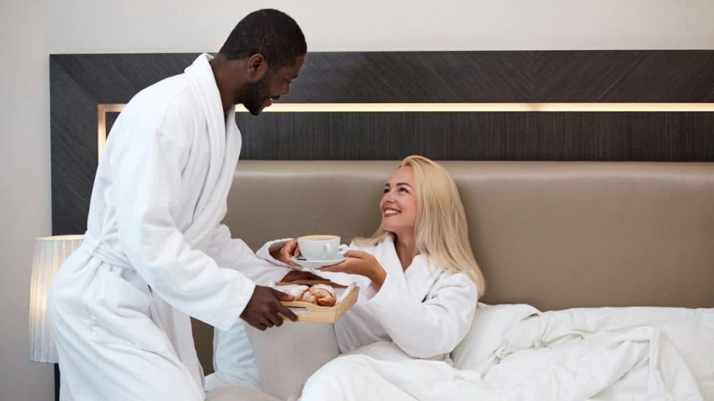 Black man in bathrobe serving breakfast in bed to a smiling blonde woman.