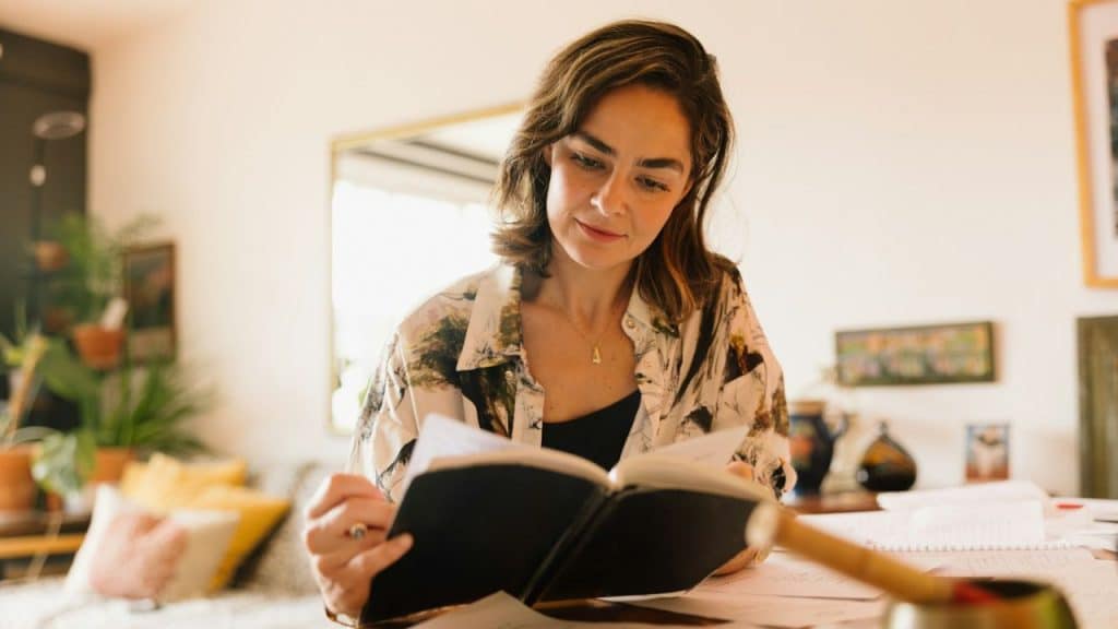 Woman wearing a patterned shirt reading an open book in a bright room.
