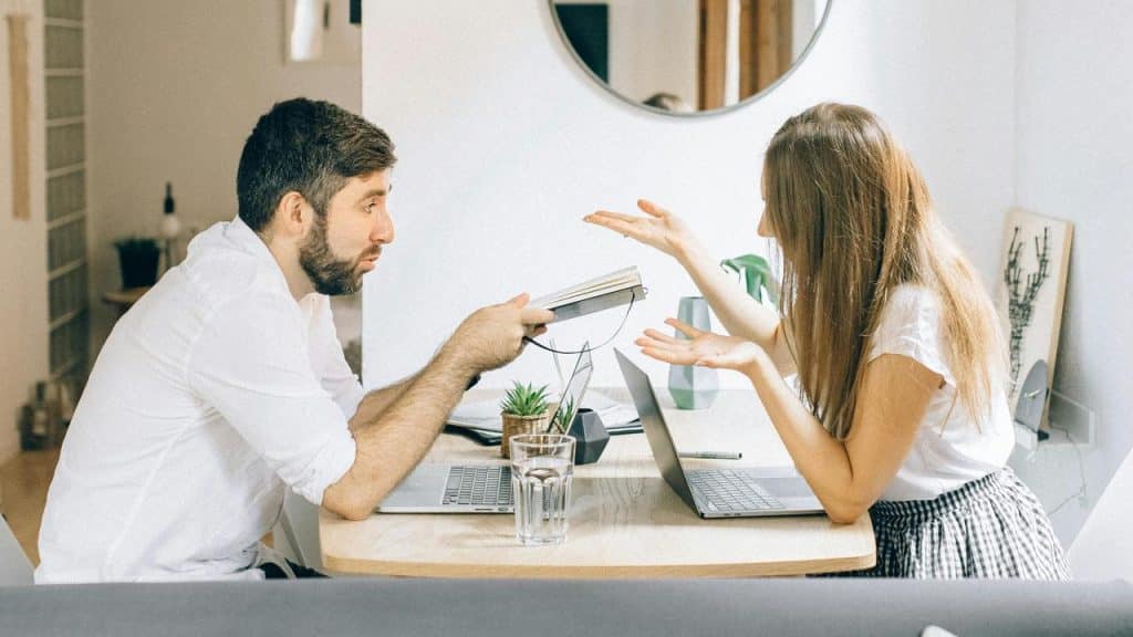A man and woman gesturing while talking across a table with laptops.