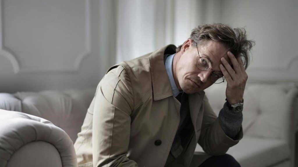 A man in glasses sitting on a sofa with his hand on his forehead.