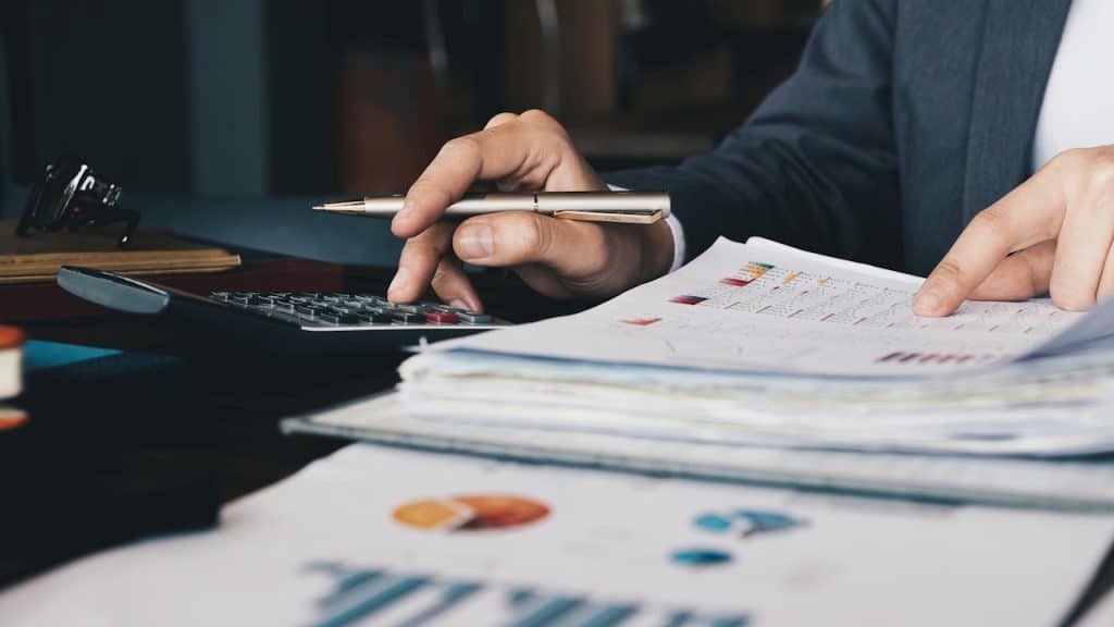 A person in a suit is using a calculator and pointing at data on a stack of financial reports.