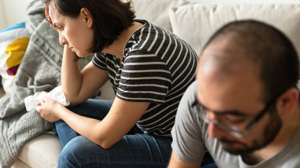 Woman sitting with a tissue, looking distressed; man in glasses out of focus in foreground.
