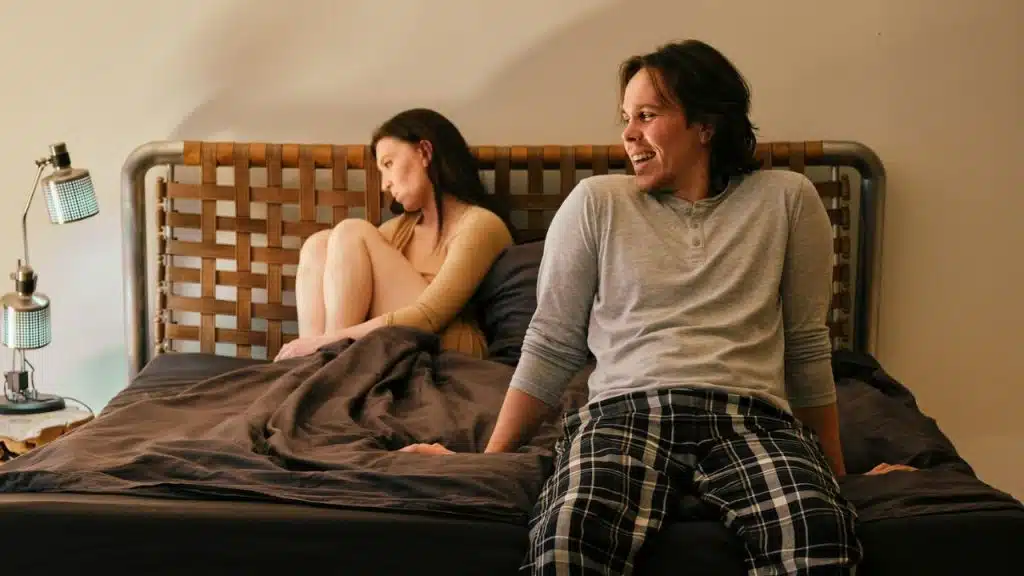 A man laughs while sitting on a bed; a displeased woman is huddled behind him.