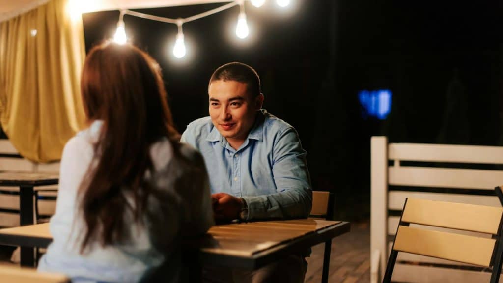Man sits across a table from a woman at night, smiling under string lights.