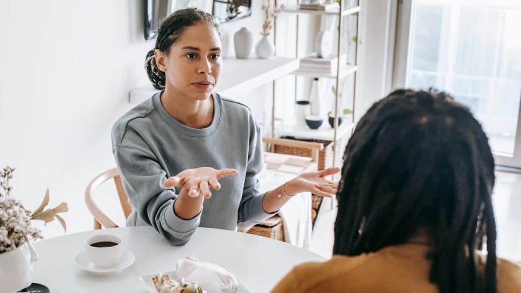 A woman with her hair pulled back gestures with her hands while talking to a person with dreadlocks across a white table.