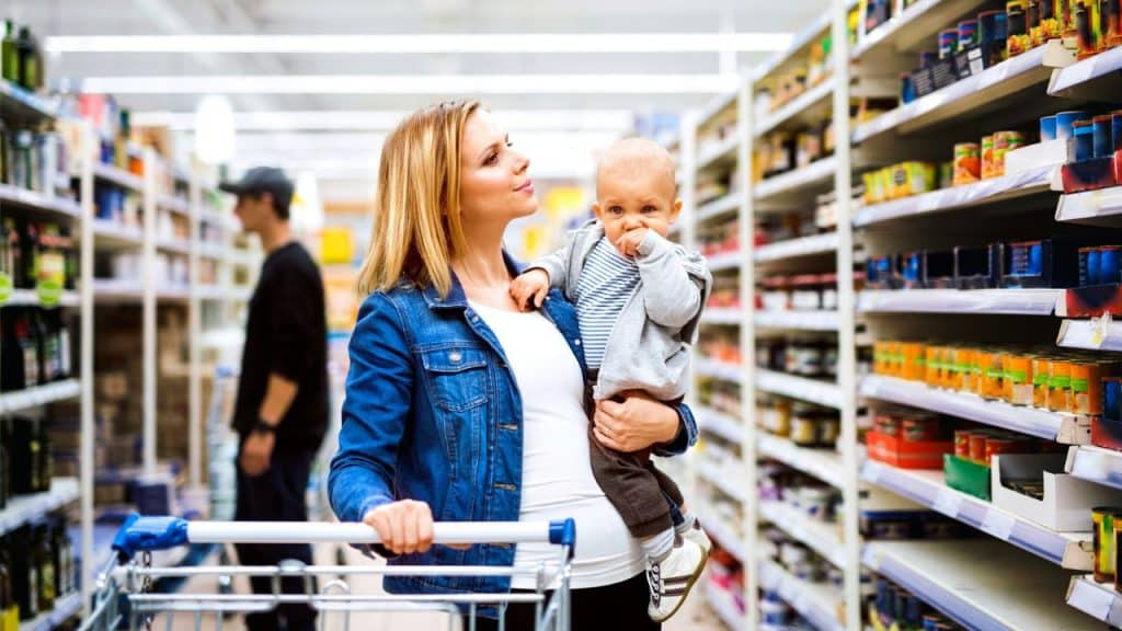 A woman holds a baby and pushes a shopping cart down a bright, wide grocery store aisle.