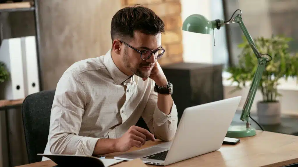 A man with dark hair and glasses is focused on his laptop while sitting at a desk with a green lamp.