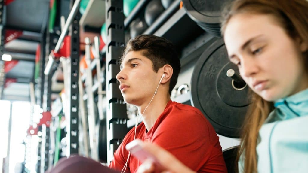 A beautiful, young, fit couple in a modern gym, resting, holding a smartphone, listening to music, and texting.