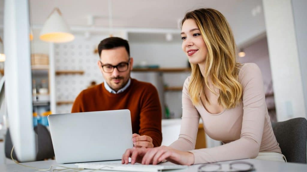 Smiling woman typing on a keyboard; man in the background works on a laptop.