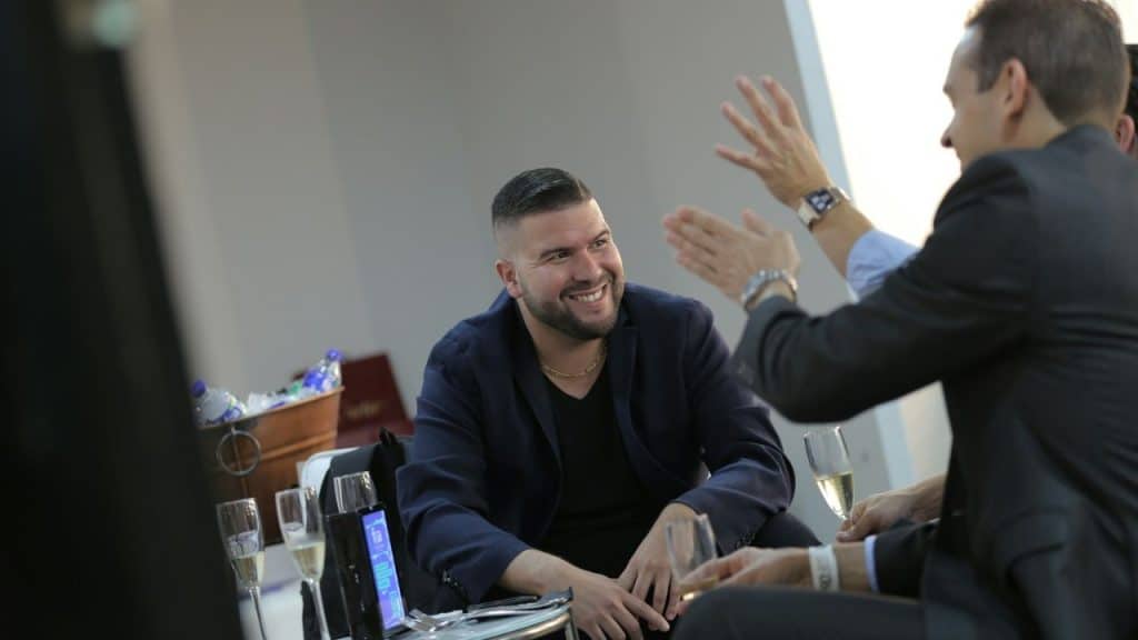 Man in dark jacket smiling while interacting with others in a modern setting.