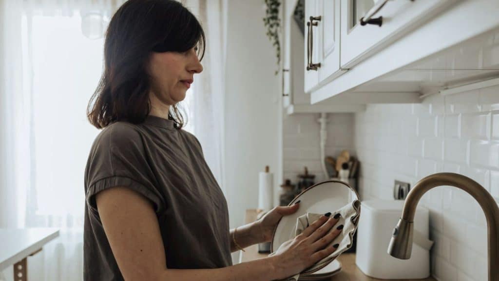 Woman in a grey shirt and black hair wiping a white plate in a kitchen.