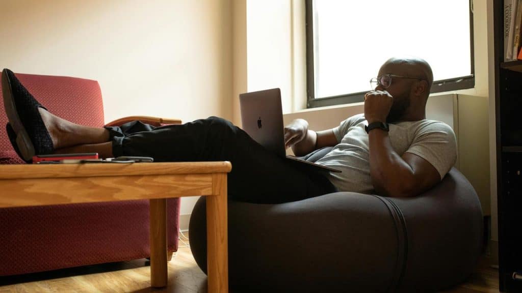 A man sitting on a bean bag using a laptop near a window.
