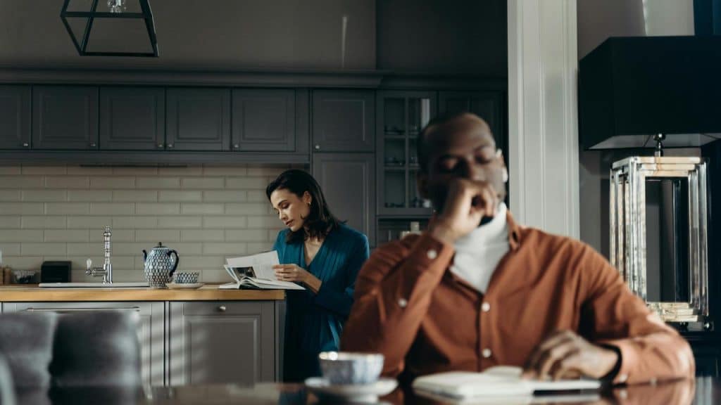 A man sitting at a table looking down while a woman reads a book in the kitchen behind him.