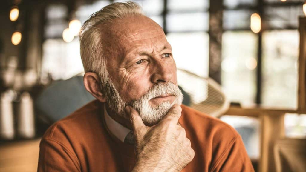 Elderly man with white beard and orange sweater holding his chin, looking away.