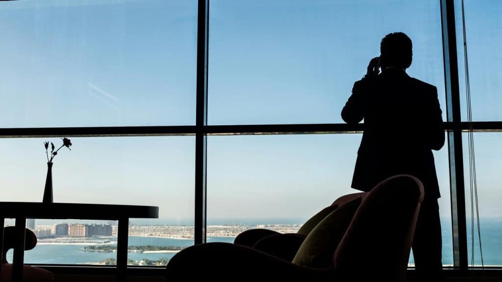 A man standing by a large window talking on the phone with a city and waterfront view.