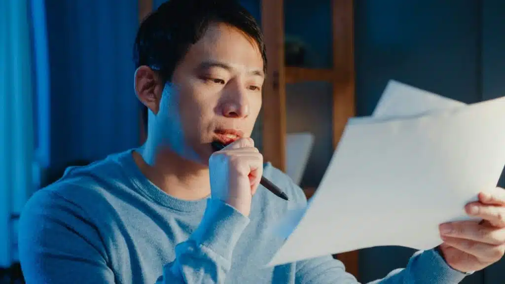 A man in a blue sweater holds papers, looking at them with a pen to his mouth.