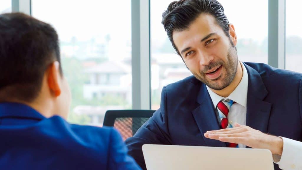 Businessman in a suit and tie leans over a laptop, talking and gesturing to colleague.