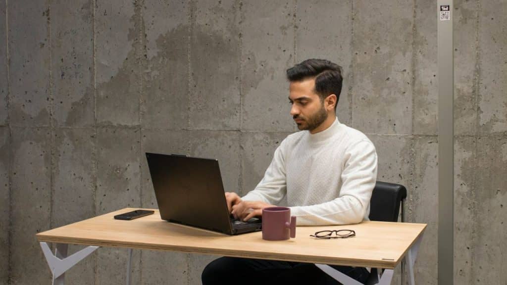 Man in a white sweater sitting at a light desk, working on a laptop.