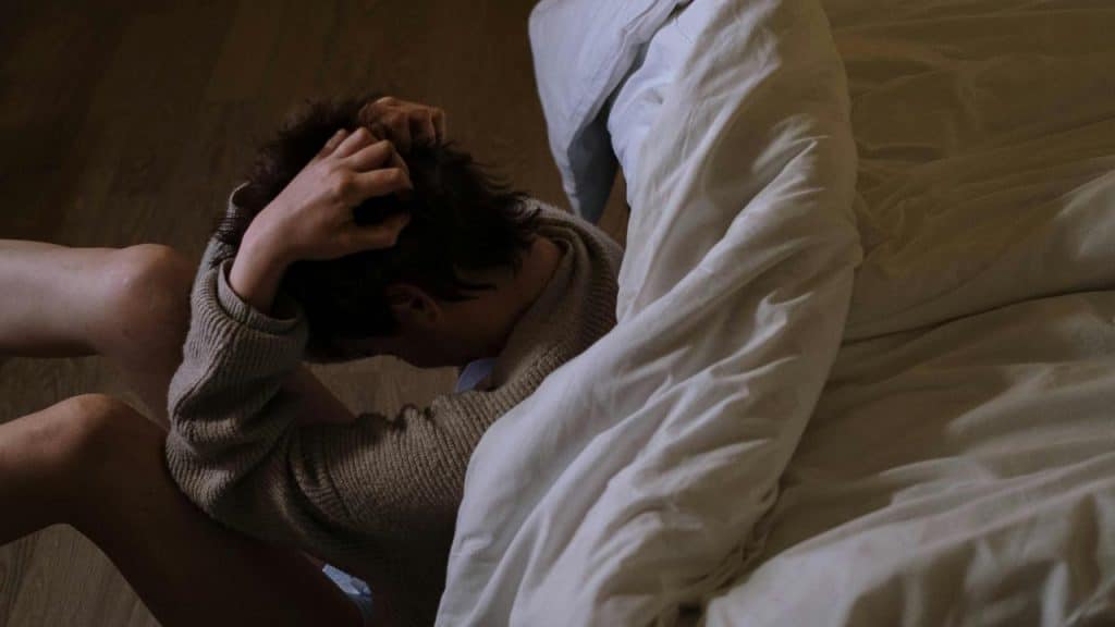 A person sits on the floor by a bed holding their head.