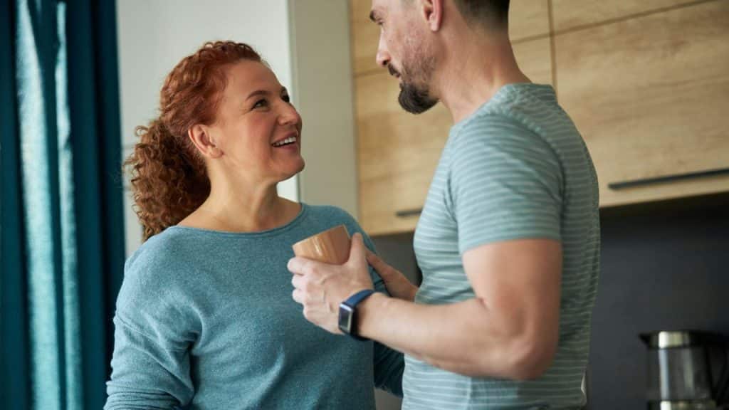 A woman with red hair smiles warmly at a man while holding a brown mug.
