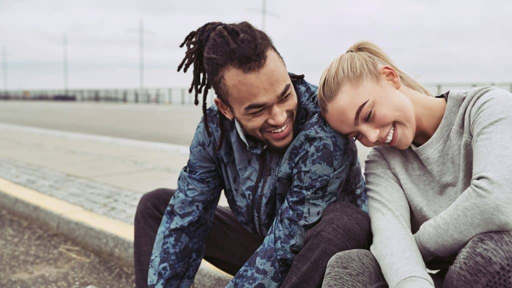 A woman resting her head on the shoulder of a man with dreadlocks as they sit on a pavement.