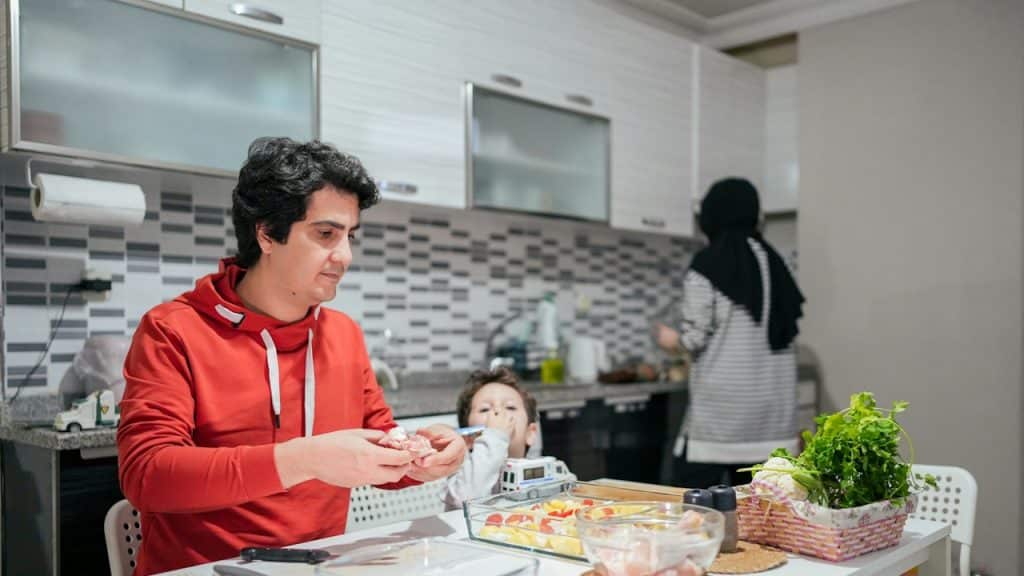 A man fixing a sandwich at a kitchen counter with his kid while a woman works in the background.