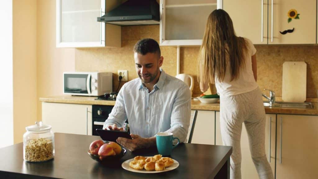 A man uses a tablet at a table while a woman works in the kitchen background.