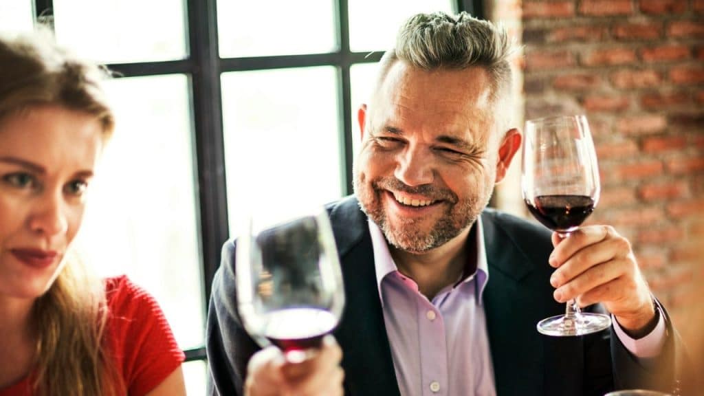 A smiling man in a suit jacket holds a glass of red wine during a toast.